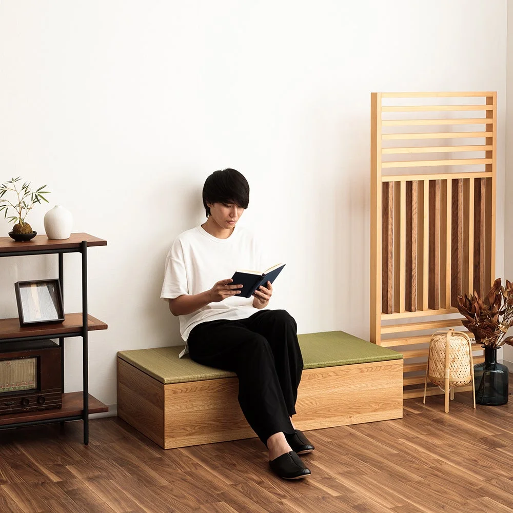 畳の収納ベンチに座って読書をする男性が写っています。和モダンな雰囲気のリビングで、木製の家具や植物が配置され、落ち着いた空間が演出されています。