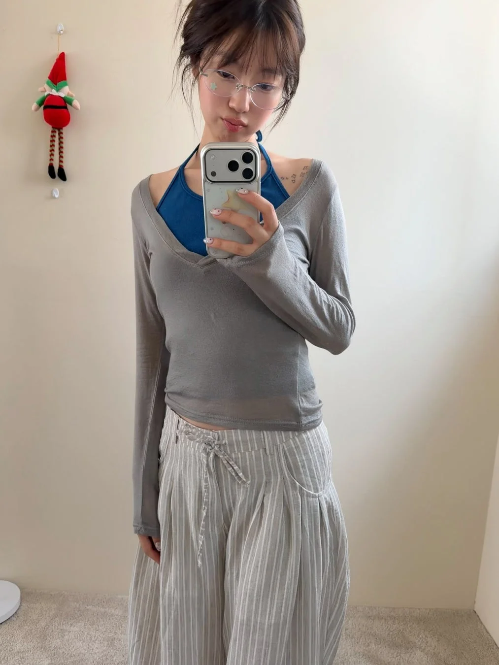 若い女性がミラーセルフィーを撮っている。グレーのシアーなトップスとストライプのワイドパンツを着用し、眼鏡をかけてスマートフォンを持っている。