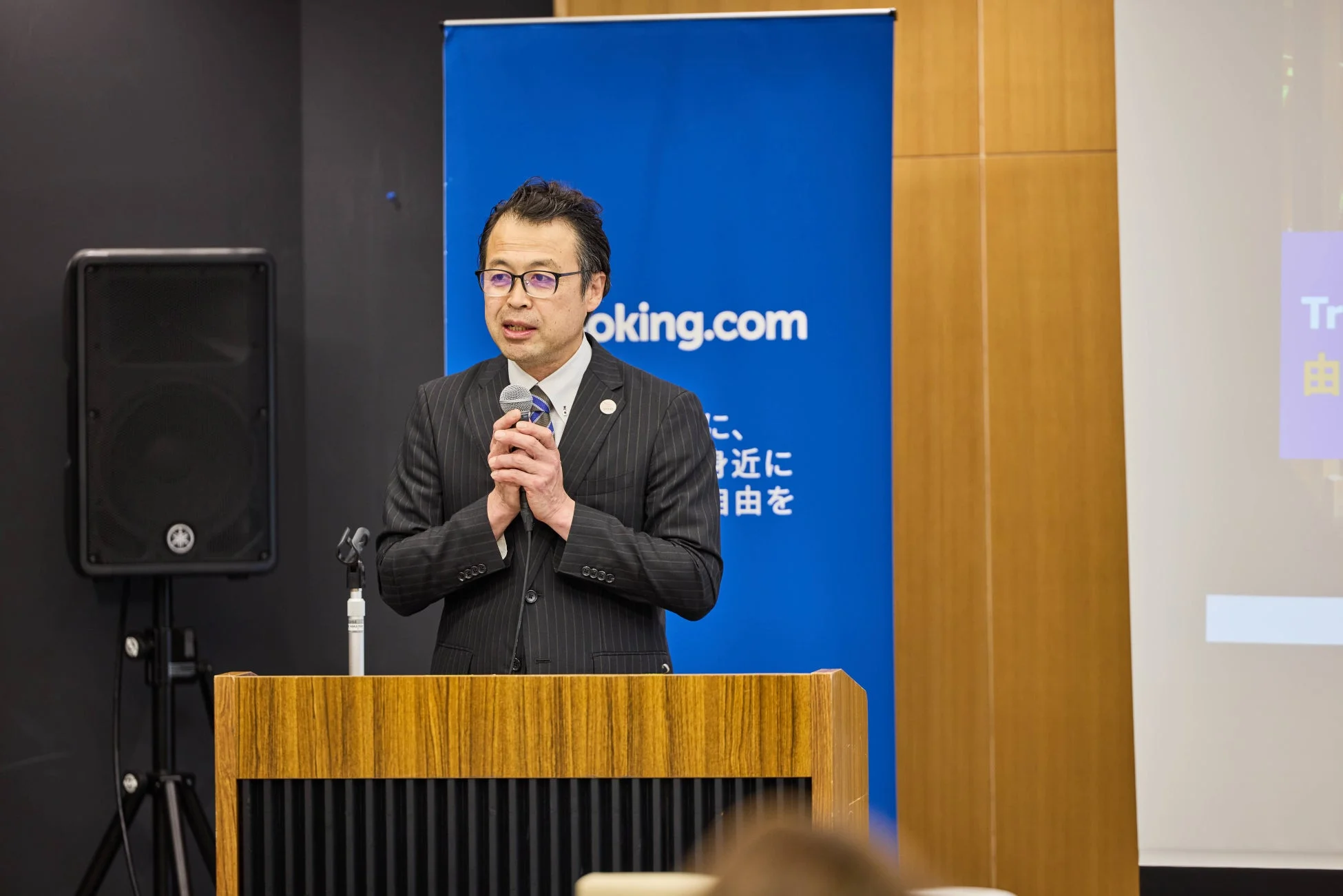 スーツ姿の男性がマイクを手に壇上で講演を行っている様子。背景には「Booking.com」のロゴが入った青いバナーと、プレゼンテーションのスライドが映し出されています。