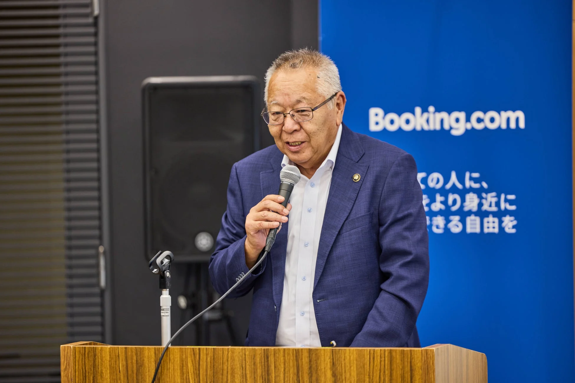 青いスーツを着た男性がマイクを持ってスピーチをしている様子。背景には「Booking.com」のロゴと「全ての人に、をより身近にできる自由を」という日本語のメッセージが書かれた青い壁が見えるビジネスイベントでの講演風景です。