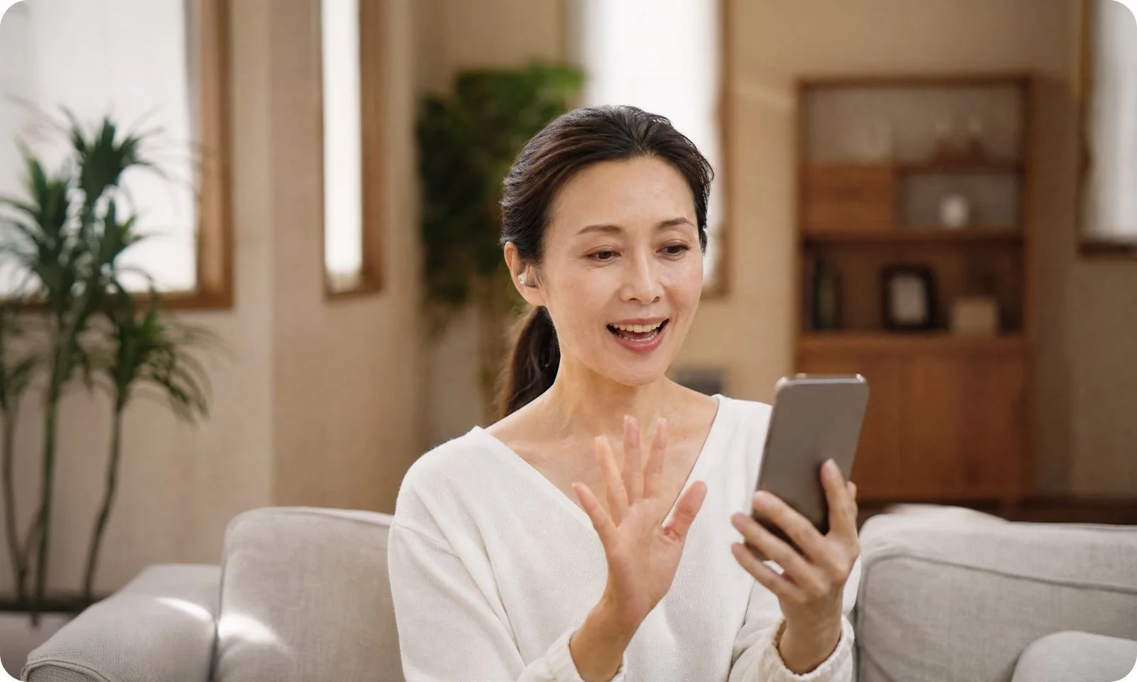 自宅のソファに座った女性が、スマートフォンを使って笑顔でビデオ通話やオンラインコミュニケーションを楽しんでいる様子です。