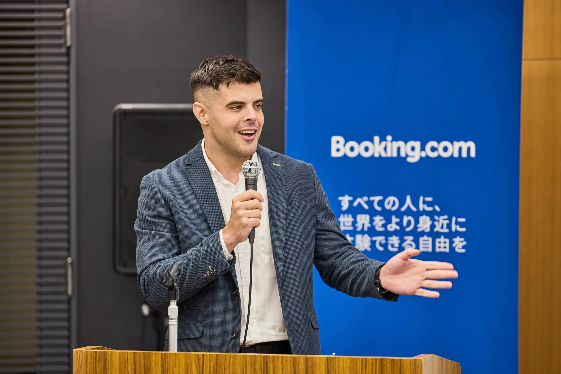 Booking.comのロゴが入った青い背景の前で、男性がマイクを手に講演を行っている様子。熱心に話しながらジェスチャーを交え、聴衆に何かを伝えている場面です。