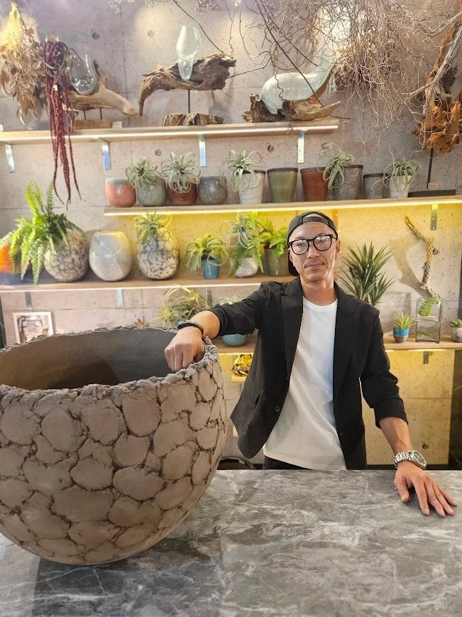 植物や鉢が並ぶ店内で、大きな個性的な鉢に手を置いた男性が写っています。背景には棚に飾られた様々な観葉植物やドライフラワーが見え、おしゃれな空間が広がっています。