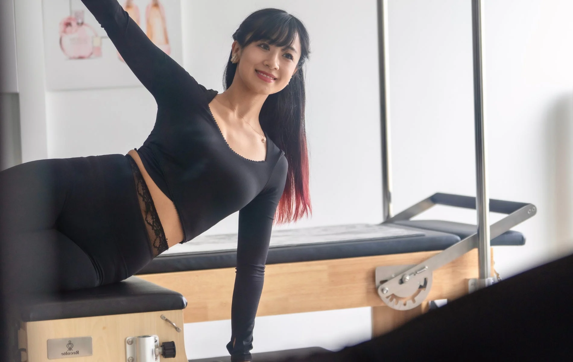 ピラティススタジオで、黒いアクティブウェアを着た女性が笑顔でピラティスリフォーマーの上でポーズをとっている。