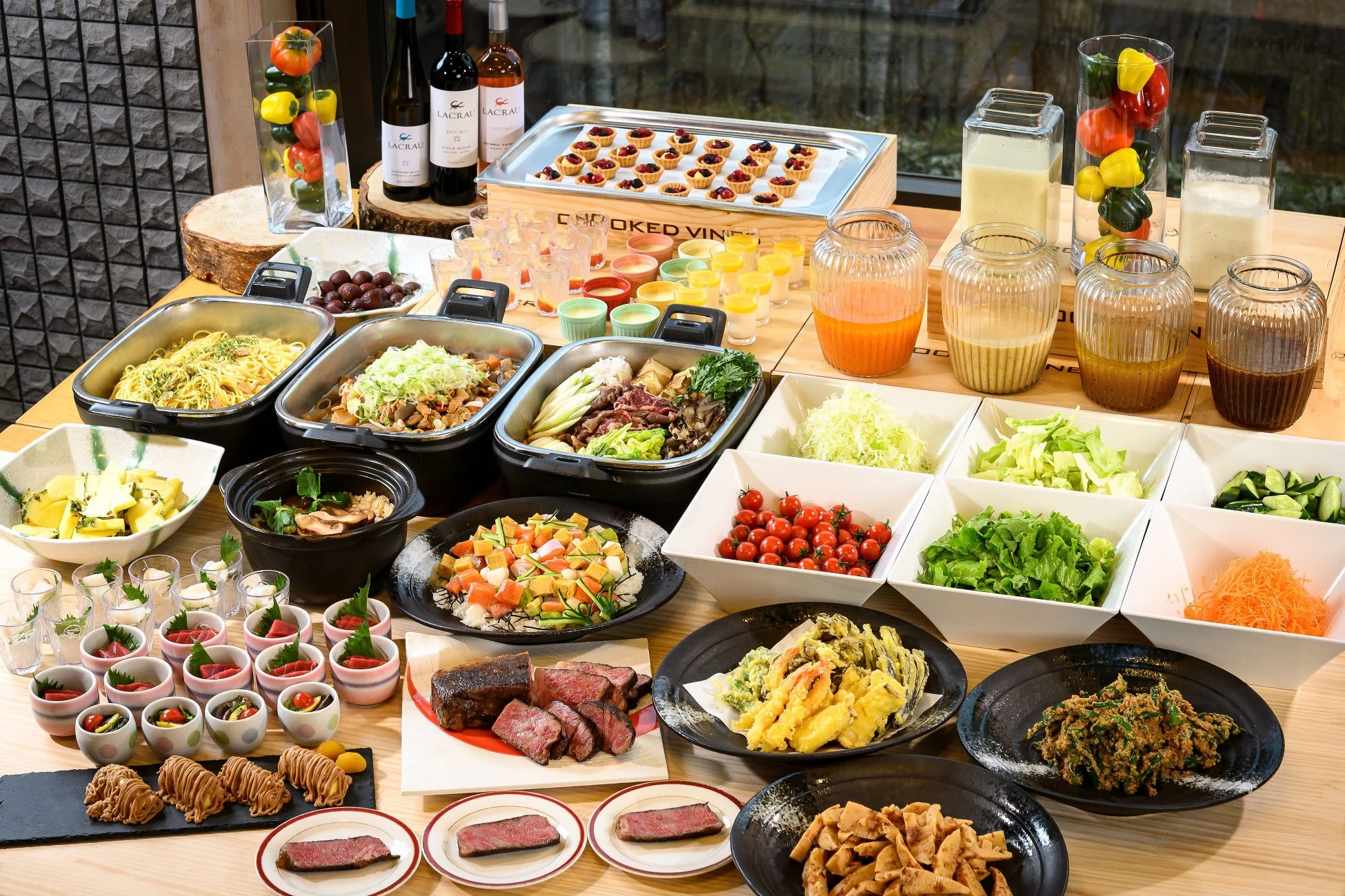 豪華なビュッフェ形式の食事が並べられたテーブルの様子。パスタや肉料理などの温かい料理、新鮮なサラダバー、天ぷら、ミニタルトなどのデザート、ワインやジュースが豊富に揃っており、多様な料理を楽しめるダイニング体験が伺えます。