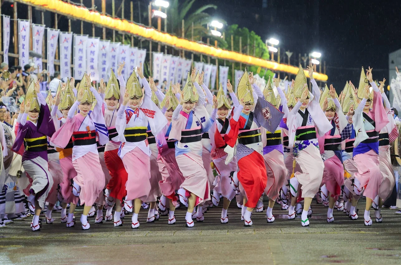 夜の祭りで、鮮やかな着物と特徴的な笠を身につけた女性たちが阿波踊りを披露しています