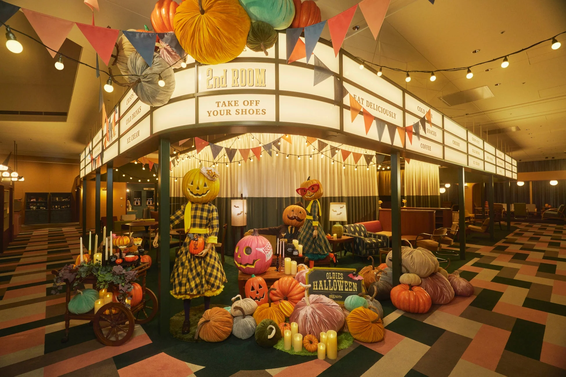 レトロな雰囲気のハロウィン装飾が施された屋内空間。カボチャの頭のキャラクター、様々なカボチャの飾り、ガーランド、電飾が賑やかな秋のイベントムードを演出しています。