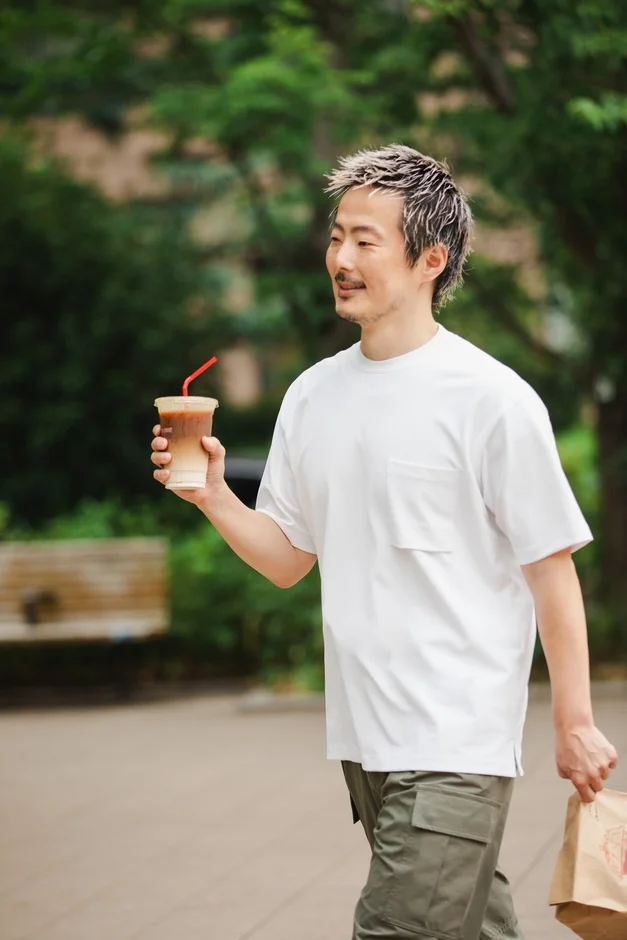 ブリーチした短髪と髭の男性が、屋外で白いTシャツとカーゴパンツを着用し、右手にアイスラテ、左手に紙袋を持って歩いています。