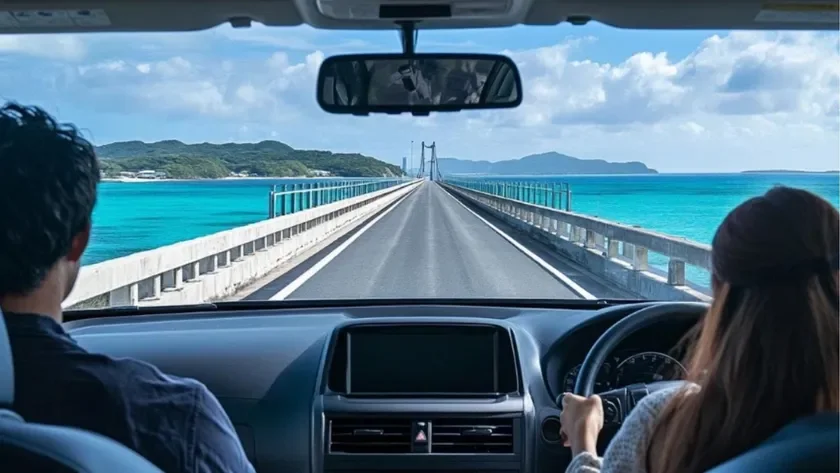 車の中から見た、青い海に架かる長い橋をドライブするカップルの様子です。晴れた空の下、エメラルドグリーンの海と遠くに見える島々が美しい絶景を形成しており、開放的な旅行気分が伝わってきます。