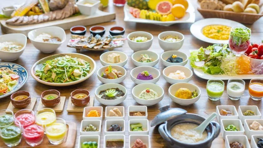 木製のテーブルに、和食を中心としたビュッフェ形式の食事が豊富に並べられています。色とりどりの小鉢料理、新鮮なサラダ、フルーツ、お粥など、健康的でバラエティ豊かな料理が美しく盛り付けられており、見た目にも楽しい食事風景です。