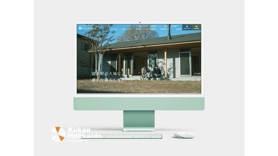 ライトグリーンのiMacが映し出されており、画面には住宅会社のウェブサイトが表示されています。ウェブサイトには、家屋の縁側に座る家族の様子と「富良野の大地に、確かな暮らしを。」というキャッチコピーが見えます。手前にはキーボードとマウスが置かれ、左下には「Kukan Hokkaido」のロゴがあります。