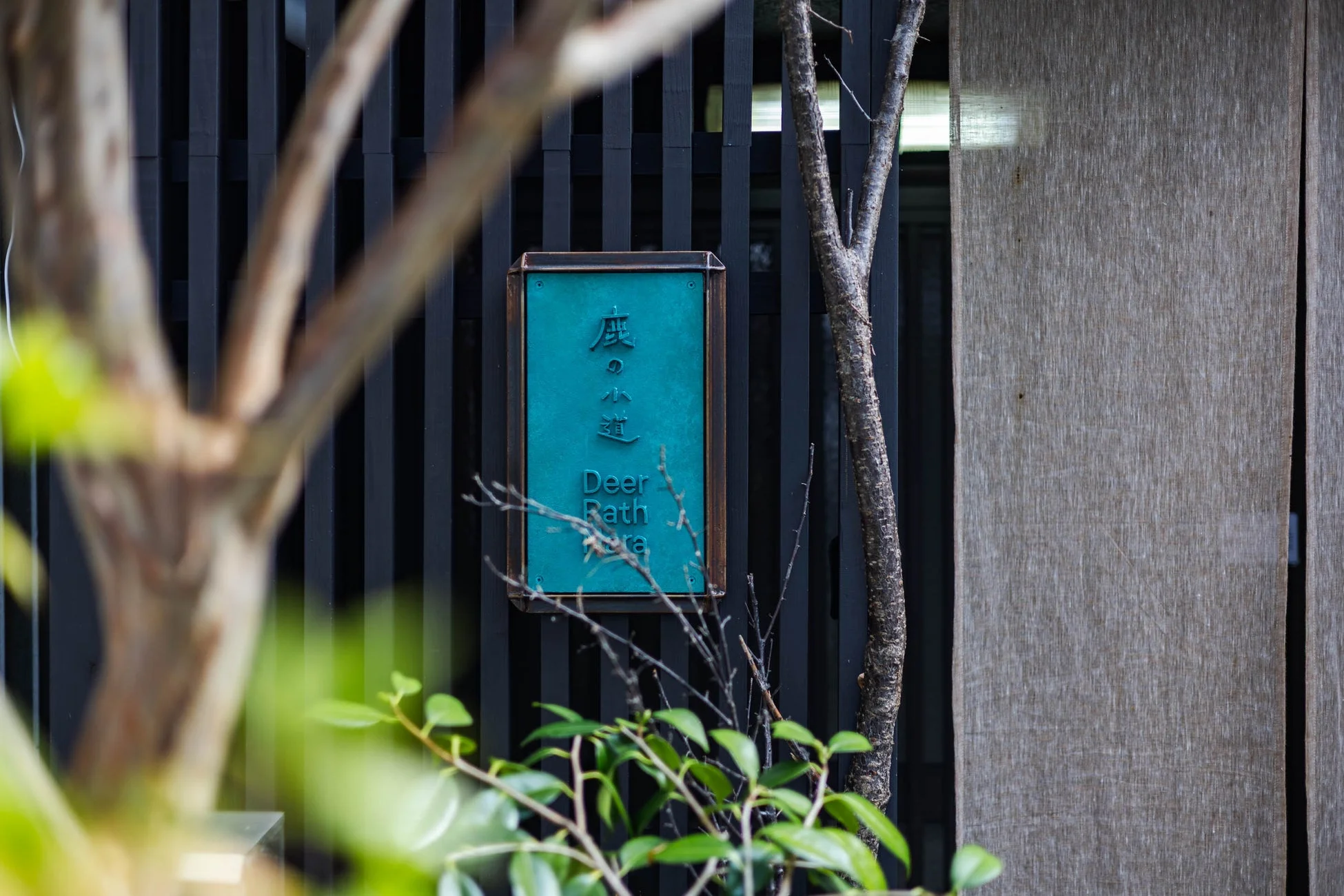 「鹿の小道」と書かれた青緑色の看板が印象的な、植物に囲まれた和風のエントランスの様子。黒い縦格子と木製の壁が落ち着いた雰囲気を醸し出しています。