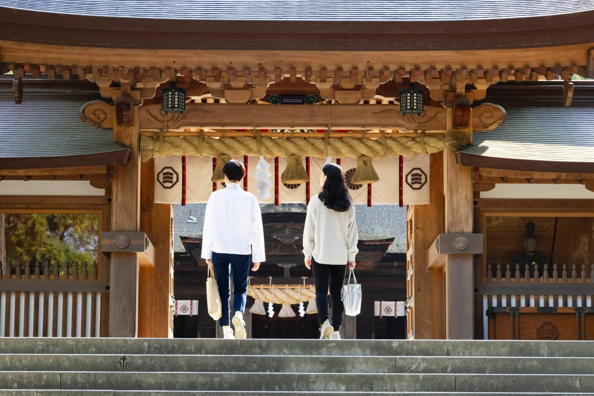 晴れた日に、二人の人物が伝統的な日本の神社建築の門をくぐろうと石段を上っている様子。しめ縄や御幣、提灯が飾られている。