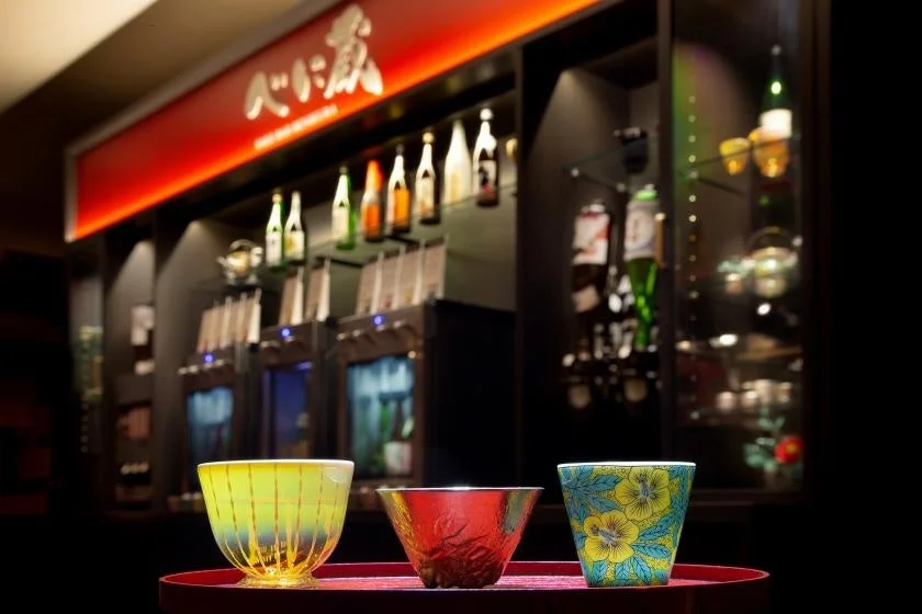 日本酒バーのような空間で、手前には色鮮やかな3つの美しい酒器が赤い盆に並べられています。