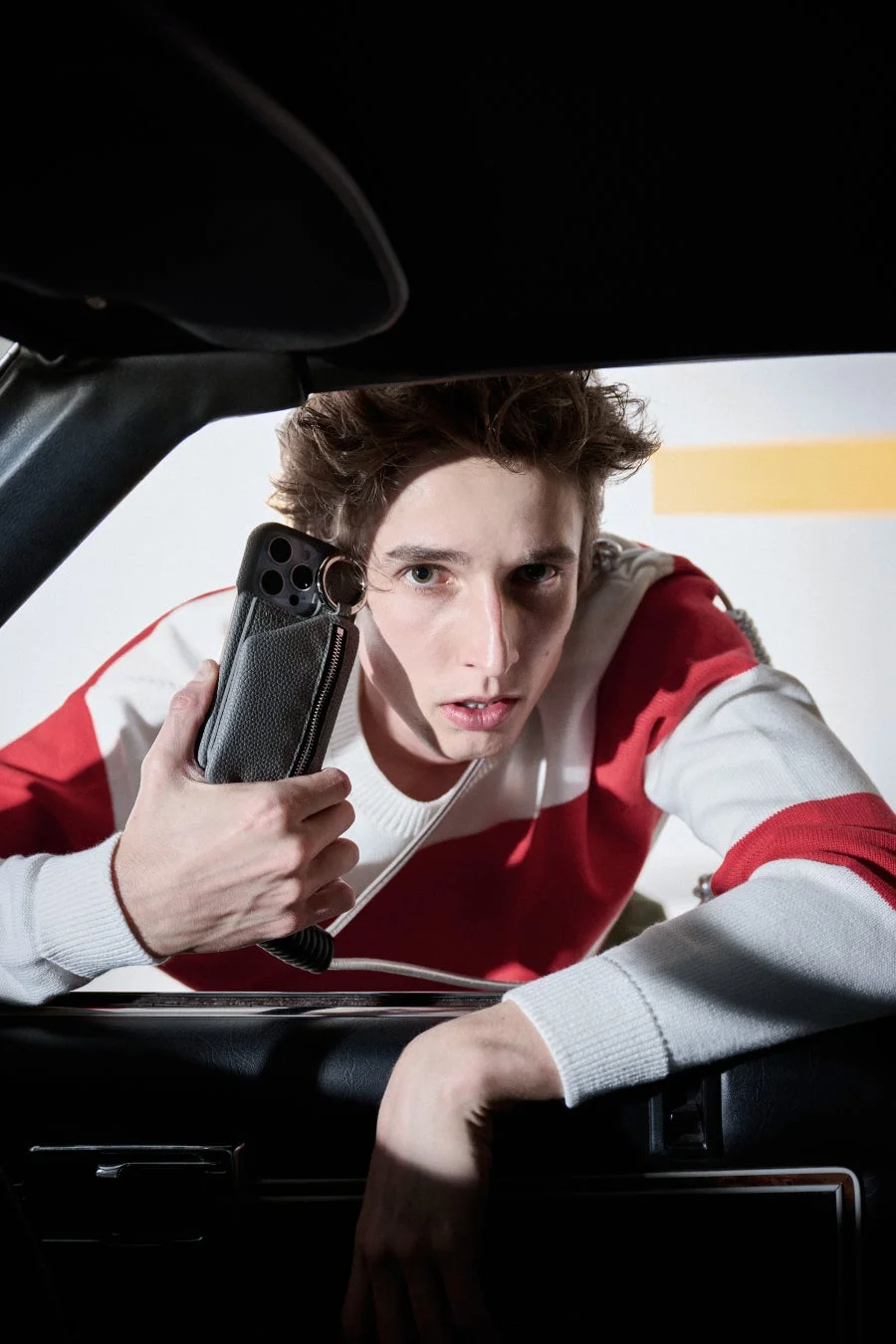 赤と白のストライプのセーターを着た若い男性が、車内のような暗い空間から身を乗り出し、黒い革のスマホケースを装着したスマートフォンを構えながらカメラをまっすぐ見つめています