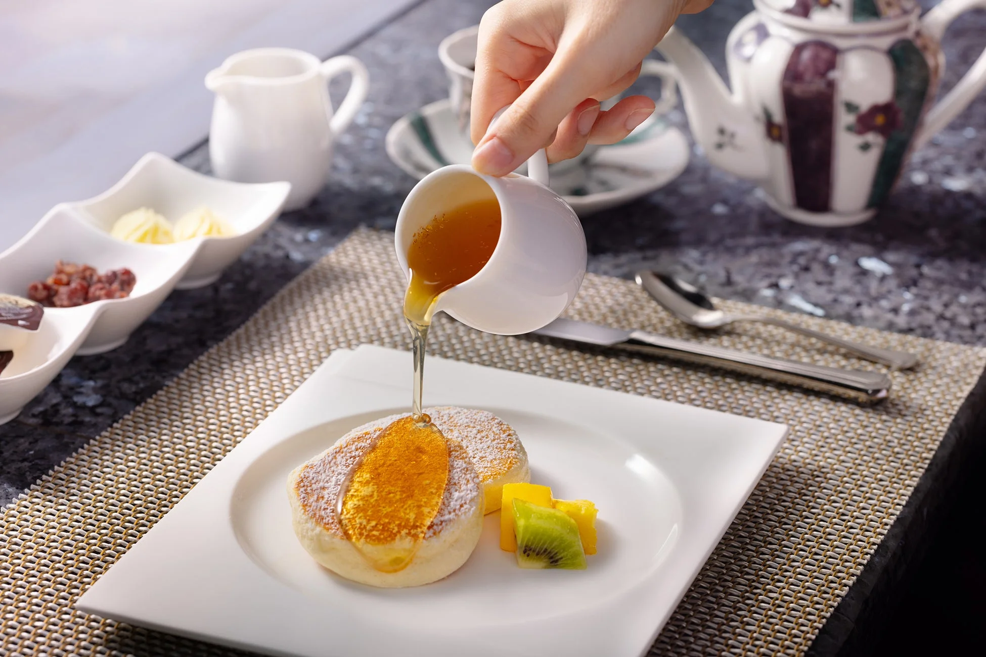 ふんわりとしたパンケーキに、白いピッチャーから黄金色のシロップがたっぷりと注がれている様子を捉えた画像です。