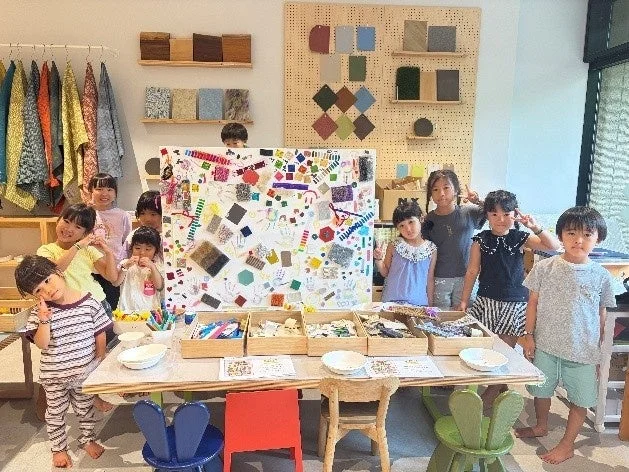 複数の子供たちがテーブルを囲み、カラフルな素材を使った共同制作を楽しんでいる様子。笑顔でカメラに向かってポーズをとっており、創造的な活動に夢中な子供たちの明るい雰囲気が伝わります。