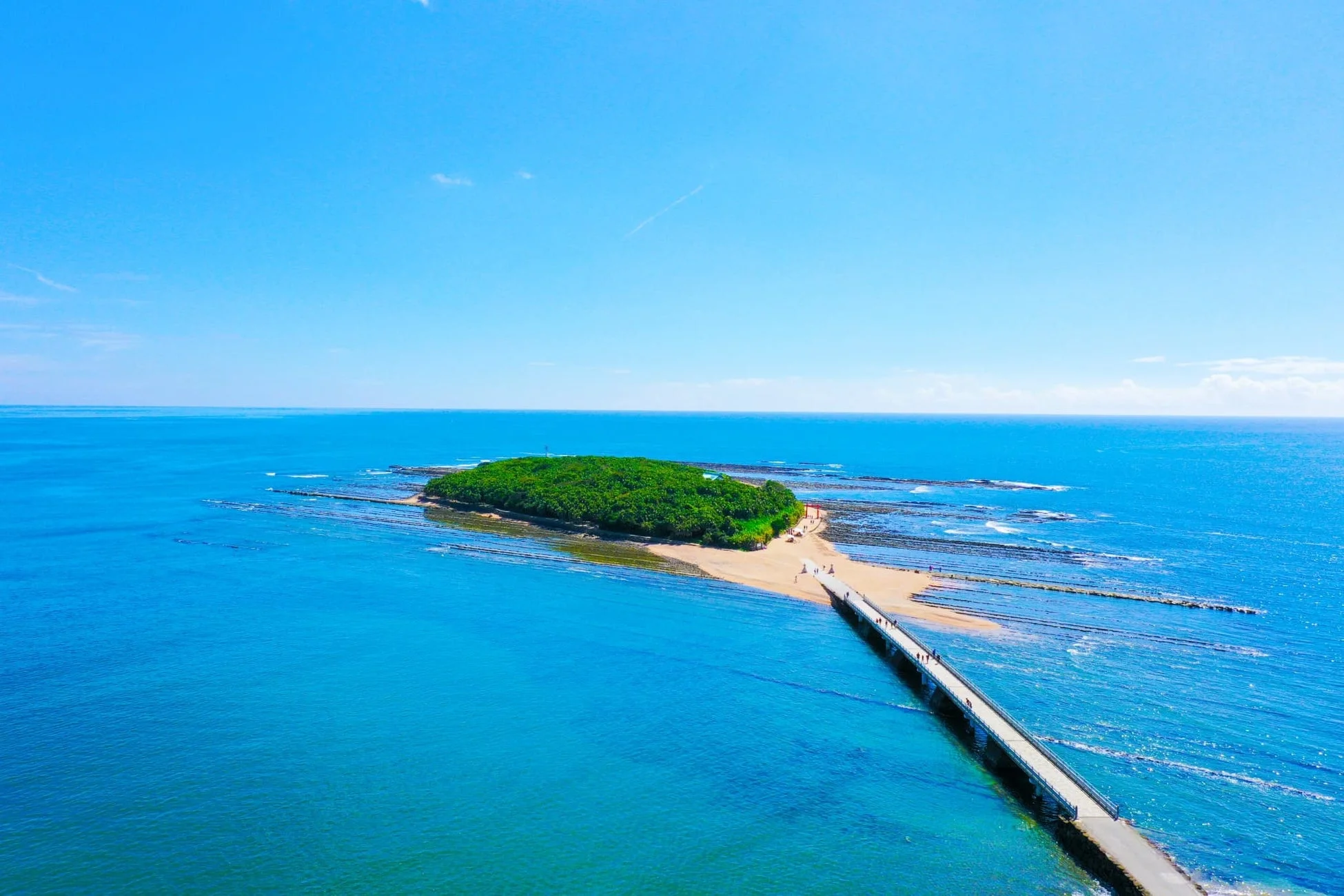 晴れ渡る青空の下、青い海に浮かぶ緑豊かな島と、そこへ続く長い橋が美しい景観を形成しています。島には鳥居が見え、橋や砂浜には多くの人が散策しており、観光地として賑わっています。