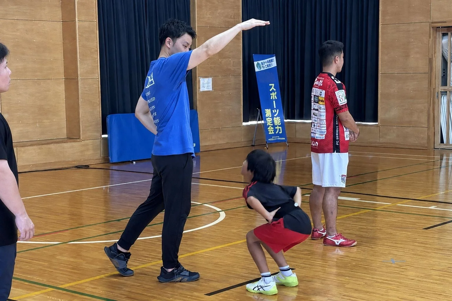 体育館で行われているスポーツ能力測定会の一幕。男性が子供に姿勢を見本として示している