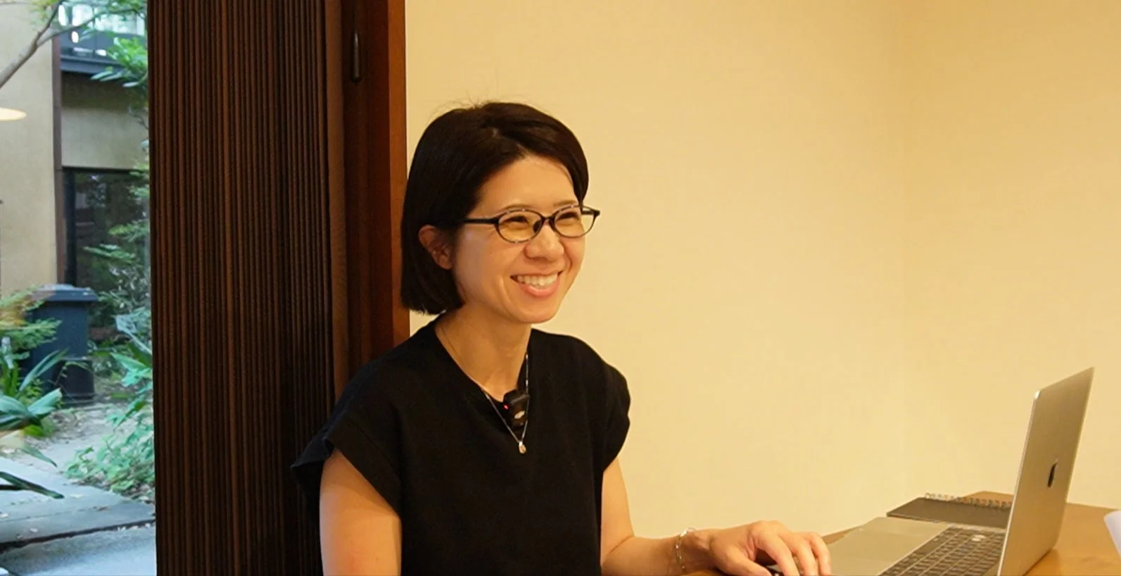 眼鏡をかけたショートヘアの女性が、ノートパソコンの前で明るく笑っています