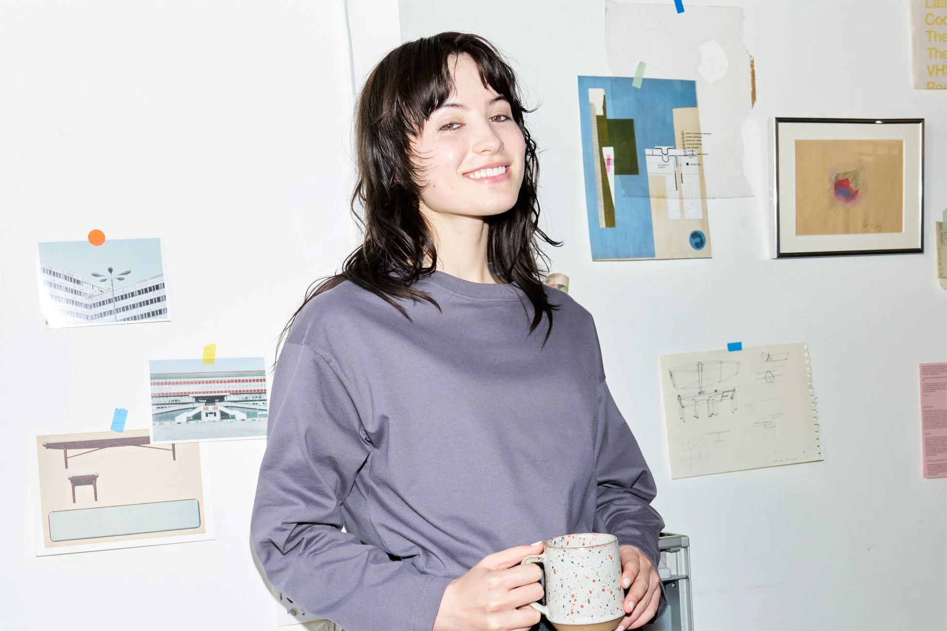 笑顔の女性がマグカップを持ち、壁にアートや写真が飾られた部屋でリラックス