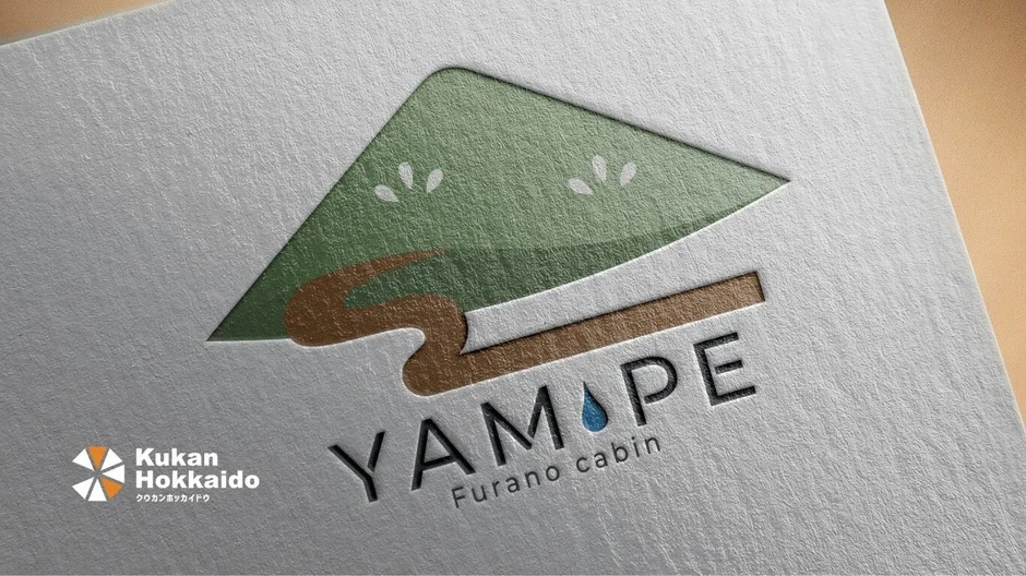 質感のある紙に「YAMAPE Furano cabin」のロゴがデザインされた画像です。ロゴは緑の山や屋根、茶色の木や道のような要素、そして「YAMAPE」の「P」に青い水滴があしらわれています。左下には「Kukan Hokkaido」のロゴも見えます。