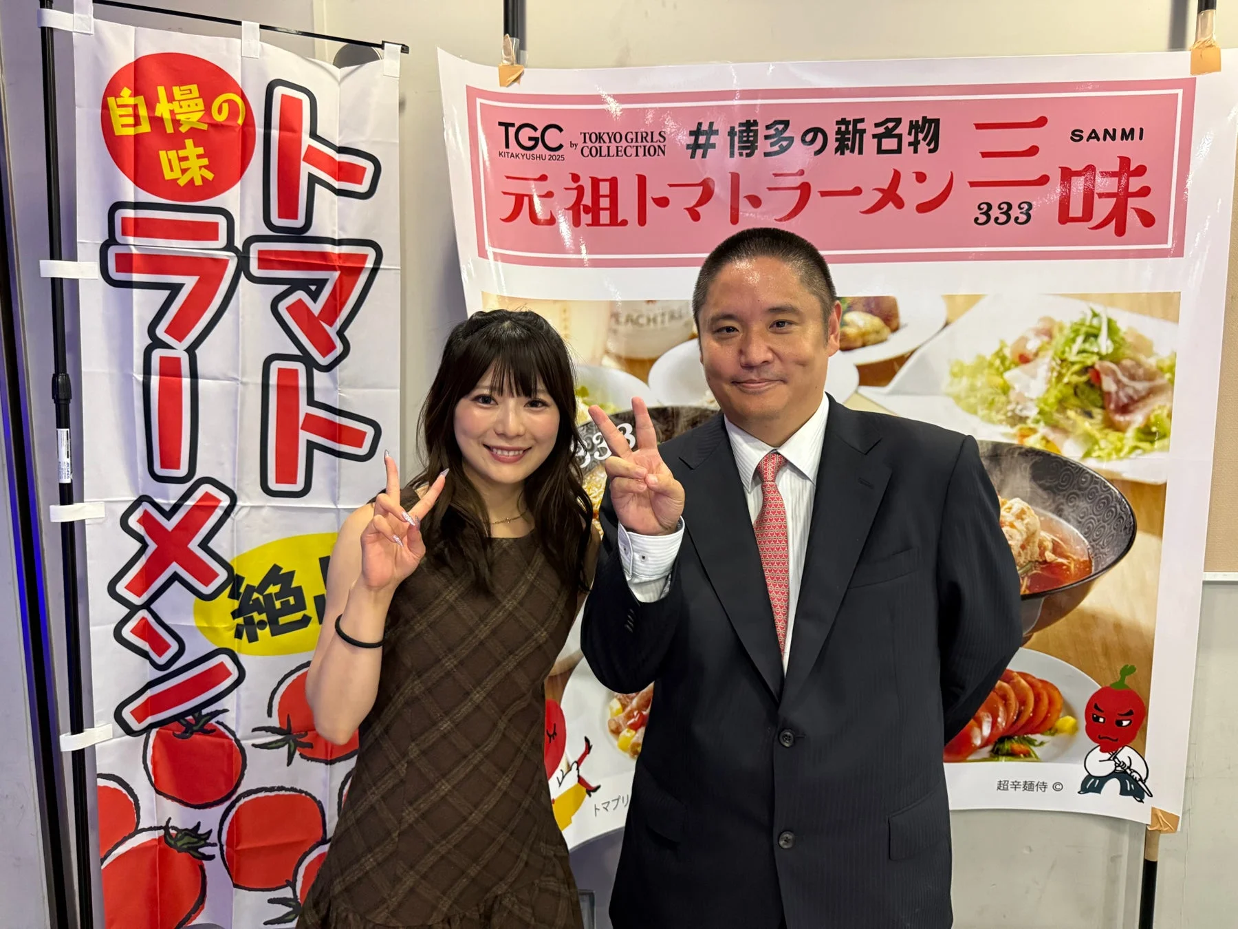 東京ガールズコレクション北九州2025のイベント会場で「元祖トマトラーメン三味」のポスターを背景に笑顔の男女