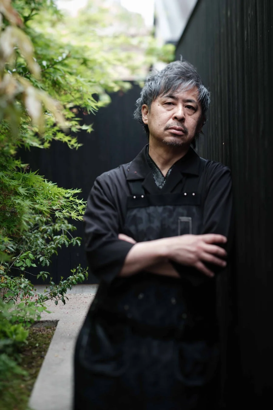 黒い服とエプロンを着用し、腕を組んで黒い壁に寄りかかる男性