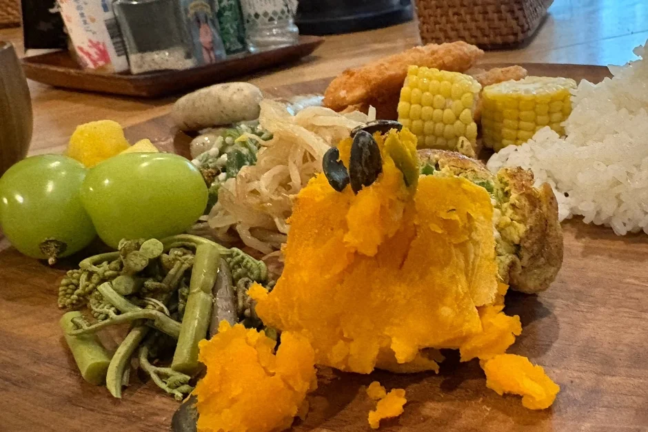 木製の皿に盛り付けられた、かぼちゃペースト、ぶどう、とうもろこし、ご飯、様々な野菜が彩り豊かに並んだヘルシーなワンプレート料理