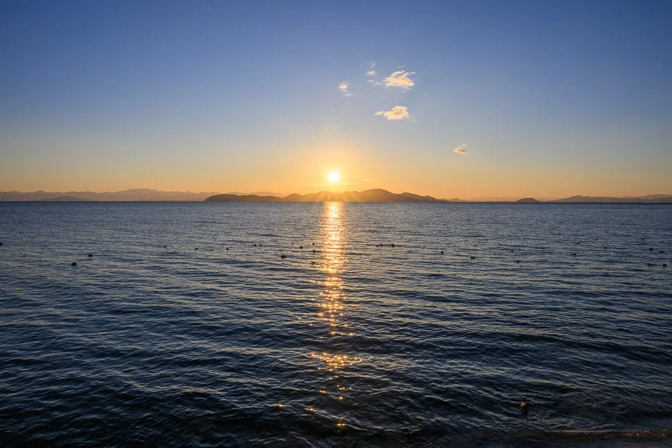夕日が水面に反射し、遠くの山々がシルエットになった美しい海の風景です。空は青からオレンジ色にグラデーションしており、穏やかな自然の瞬間を捉えています。