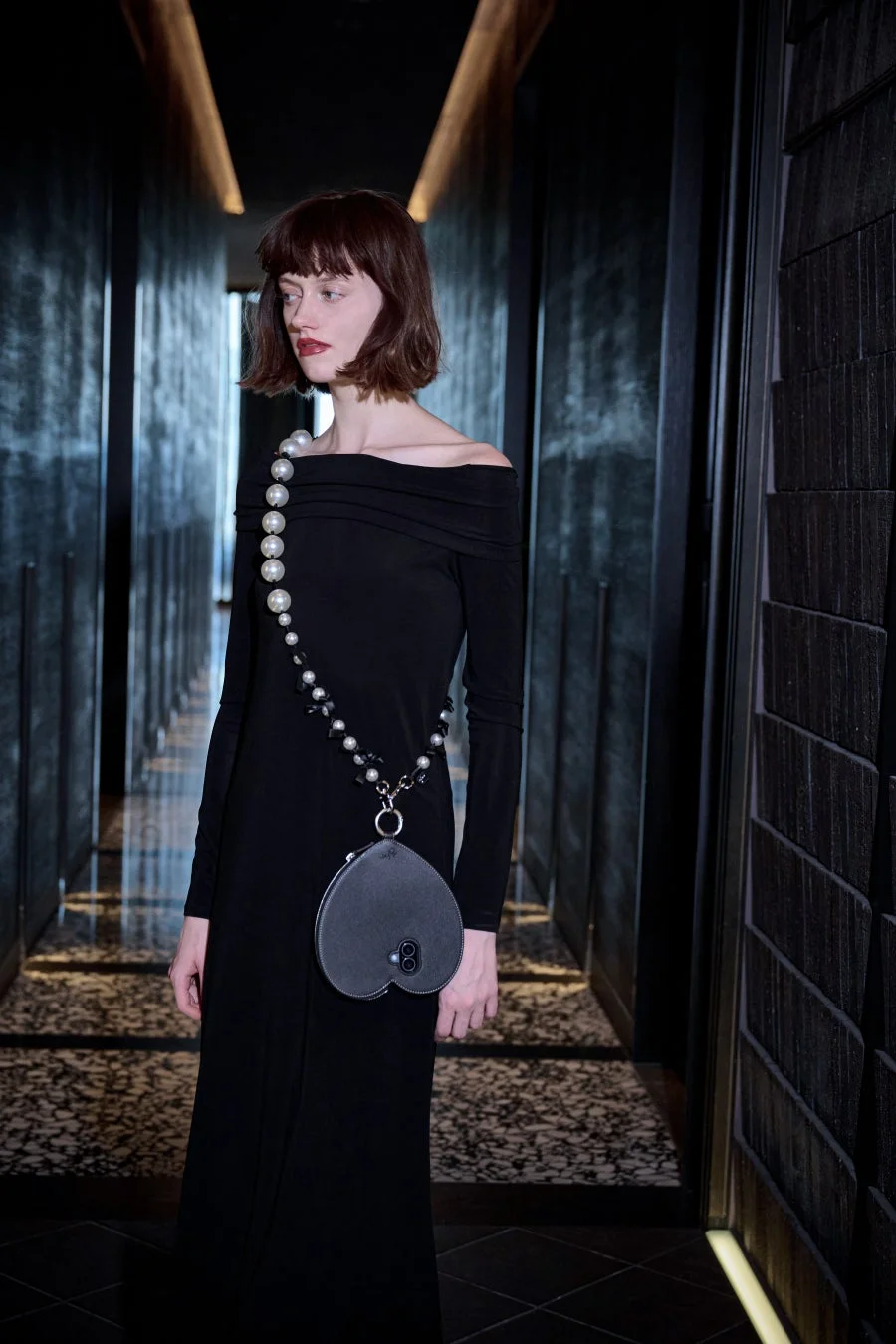 暗い廊下に立つ女性のファッションポートレート。オフショルダーの黒いロングドレスを着用し、パールと黒い飾りのストラップが付いたハート型のバッグを身につけている