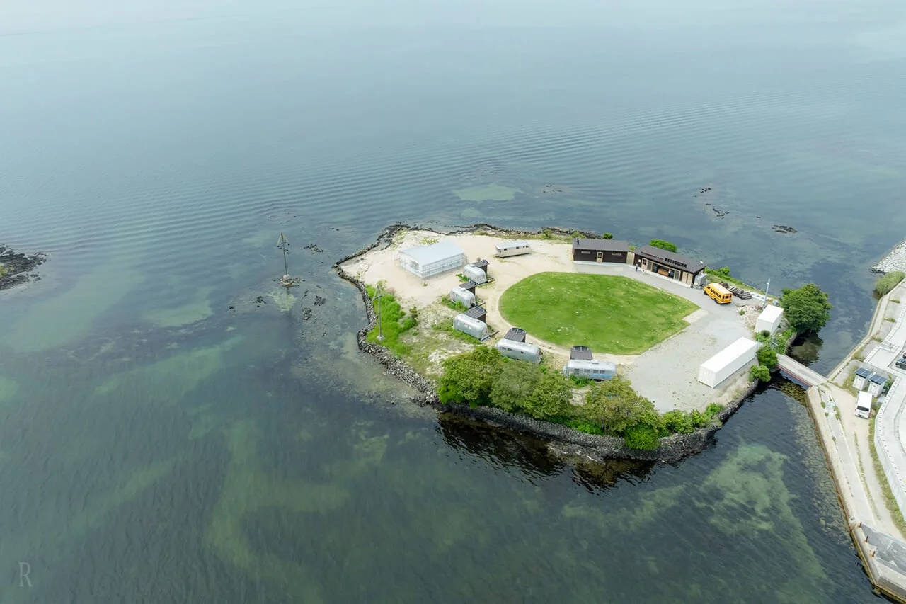 水辺に浮かぶ小さな半島を空撮した画像。複数のトレーラーハウス、建物、緑の広場、黄色いバスが見られる。