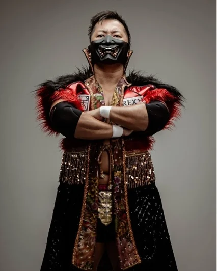 鬼のような黒いマスクと豪華な衣装を身につけた男性が腕を組み、正面を見据えているポートレート。赤いファーと金色の装飾が特徴的な衣装で、力強い印象を与えます。