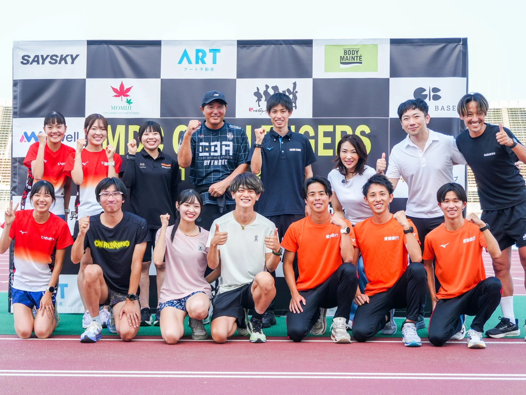 陸上競技場で撮影された、男女アスリートと関係者による集合写真。スポンサーロゴ入りの背景の前で、皆が笑顔で拳を上げたり親指を立てたりして、意気込みを示しています。