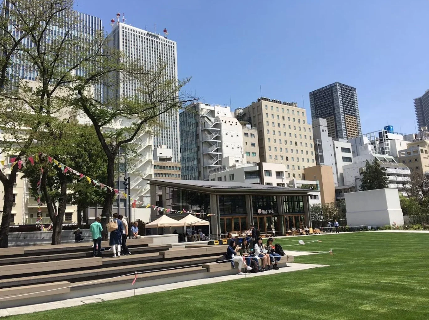 都会のビル群を背景に、広々とした芝生の公園が広がる風景です。カフェの前に人々が集まり、階段状のベンチに座ってくつろいでいます。晴れた日の開放的な都市空間が感じられます。