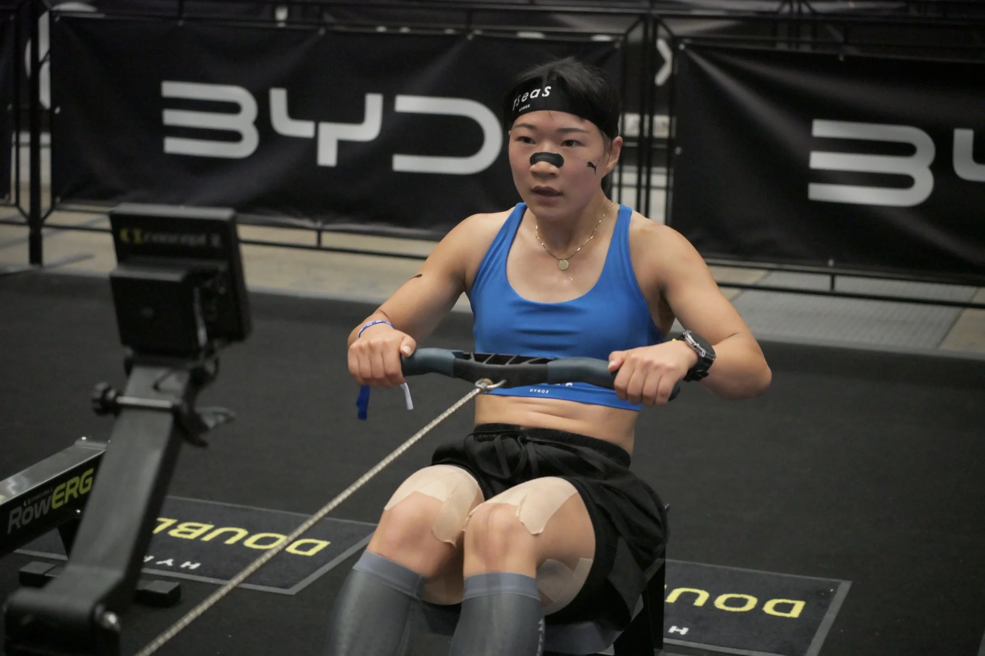 ローイングマシンで真剣に運動する女性アスリート。青いスポーツブラを着用し、鼻と膝にはテーピングが見られます。HYROXのロゴが入ったヘッドバンドをつけ、集中した表情でトレーニングに励んでいます。