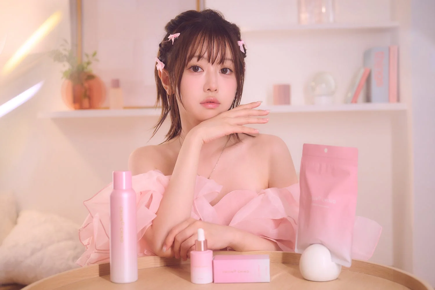 ピンクのオフショルダートップを着た女性が、複数のピンク色のスキンケア製品と一緒にポーズをとっています。