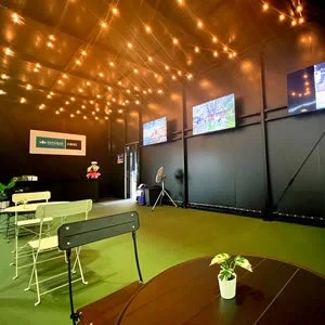 暗い天井と壁にストリングライトが輝く屋内のエンターテイメント施設です。緑の床と複数の大型スクリーンが設置されており、ゴルフシミュレーターやゲームが楽しめるラウンジのような空間が広がっています。手前にはテーブルと椅子が配置されています。