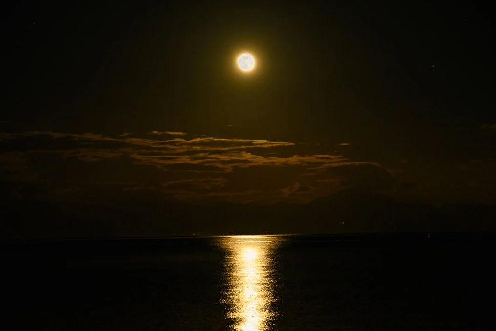 暗い夜空に輝く満月が、広がる水面に明るい光の道を映し出しています。いくつかの雲が月明かりに照らされ、幻想的で静かな夜の海の風景を作り出しています。