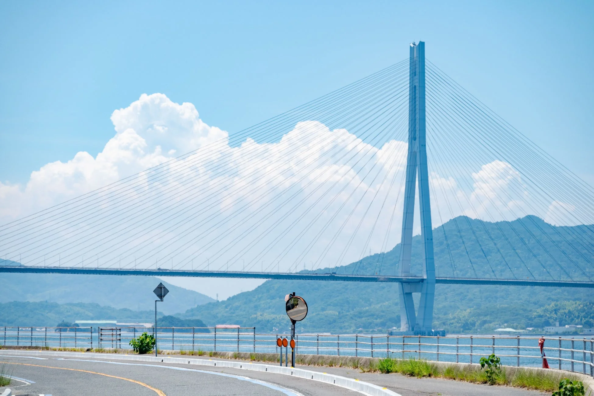 青い空と白い雲の下、広大な海に架かる雄大な斜張橋の風景です。手前にはカーブした道路とガードレールがあり、遠くには緑豊かな山々が広がっています。