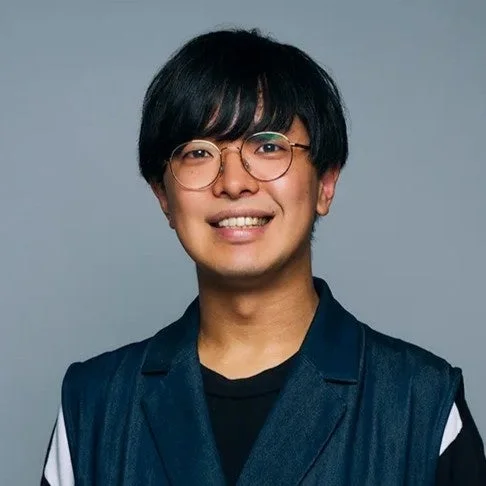 眼鏡をかけた黒髪の若いアジア人男性が、グレーの背景の前で笑顔を見せています。ダークブルーのベストを着用しており、親しみやすい印象を与えます。