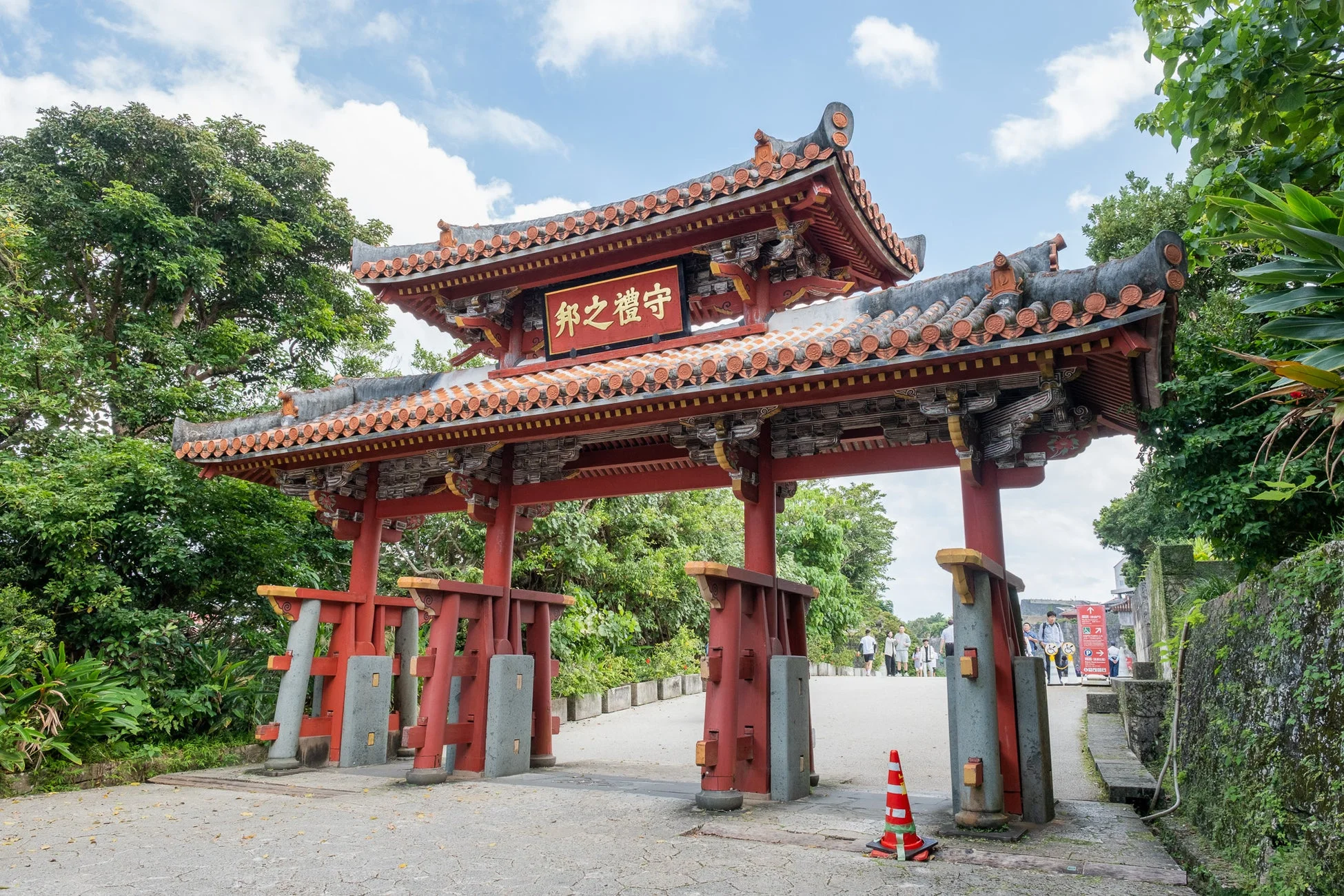 沖縄の首里城公園にある象徴的な守礼門が、青空と緑豊かな木々を背景に写っています。赤い瓦屋根と柱が特徴的な伝統的な建築様式で、門の上部には「守禮之邦」の文字が見えます。