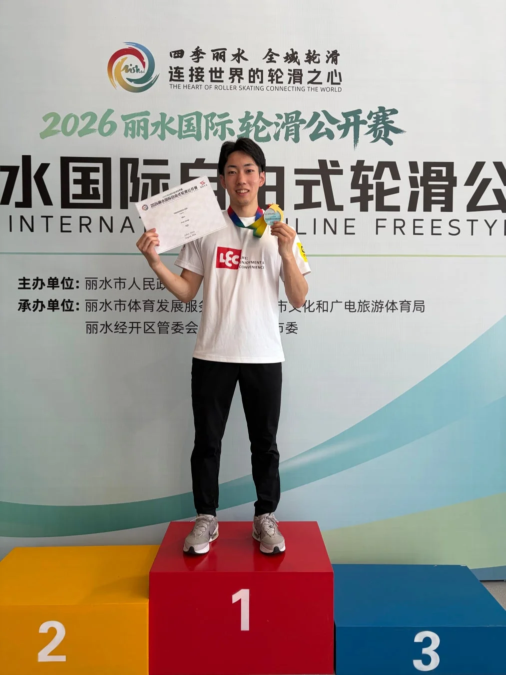 2026年麗水国際輪滑公開賽の表彰式で、金メダルと賞状を手に笑顔で表彰台の1位に立つ男子選手