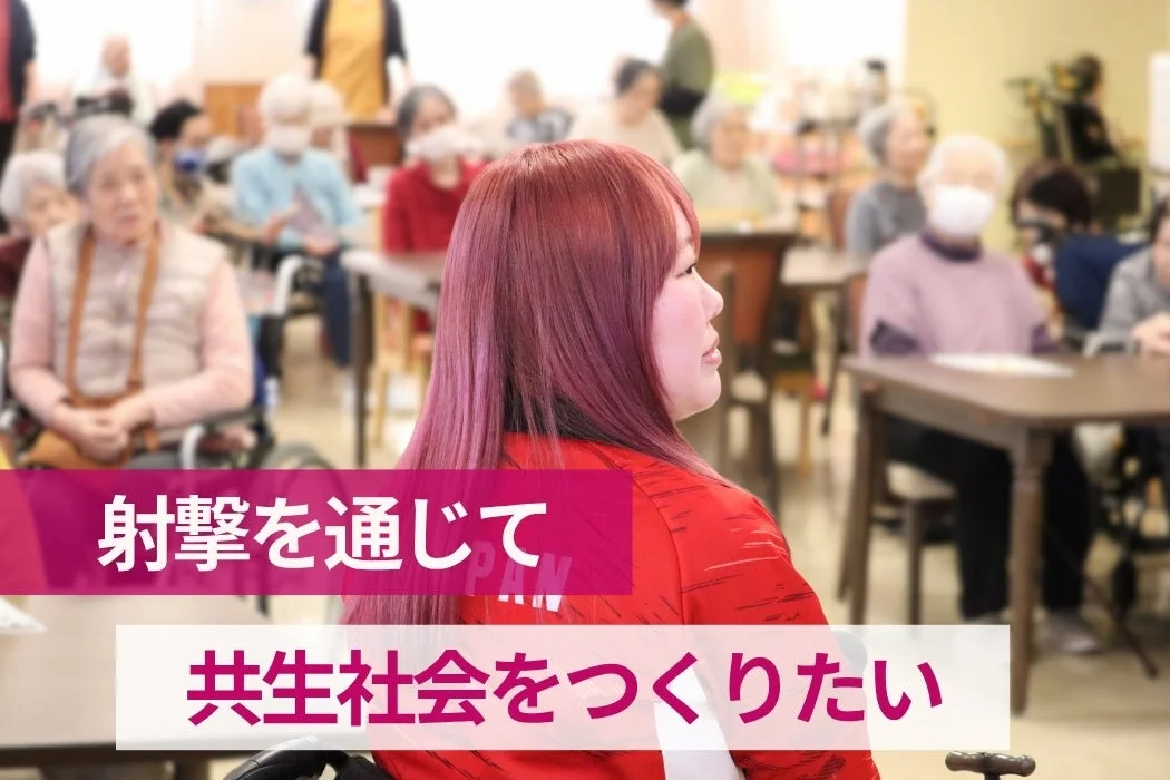 赤いジャケットを着た女性が、多くの高齢者が集まる室内で座っている画像