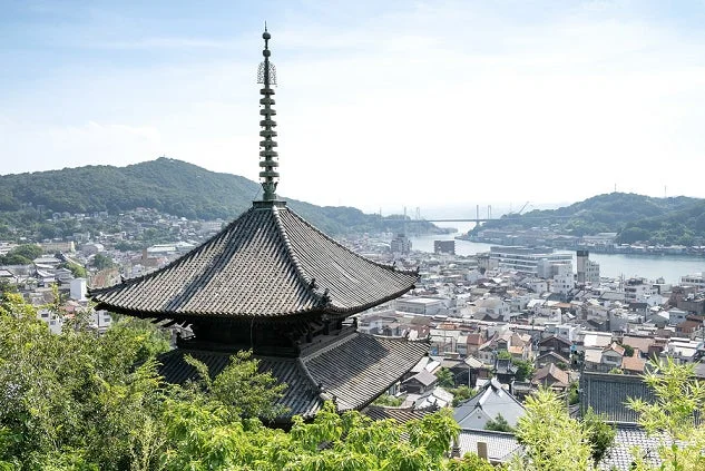 日本の伝統的な仏塔が前景にそびえ立ち、その下には海と橋が見える美しい港町の全景が広がっている。緑豊かな山々に囲まれた、晴れた日の日本の風景。