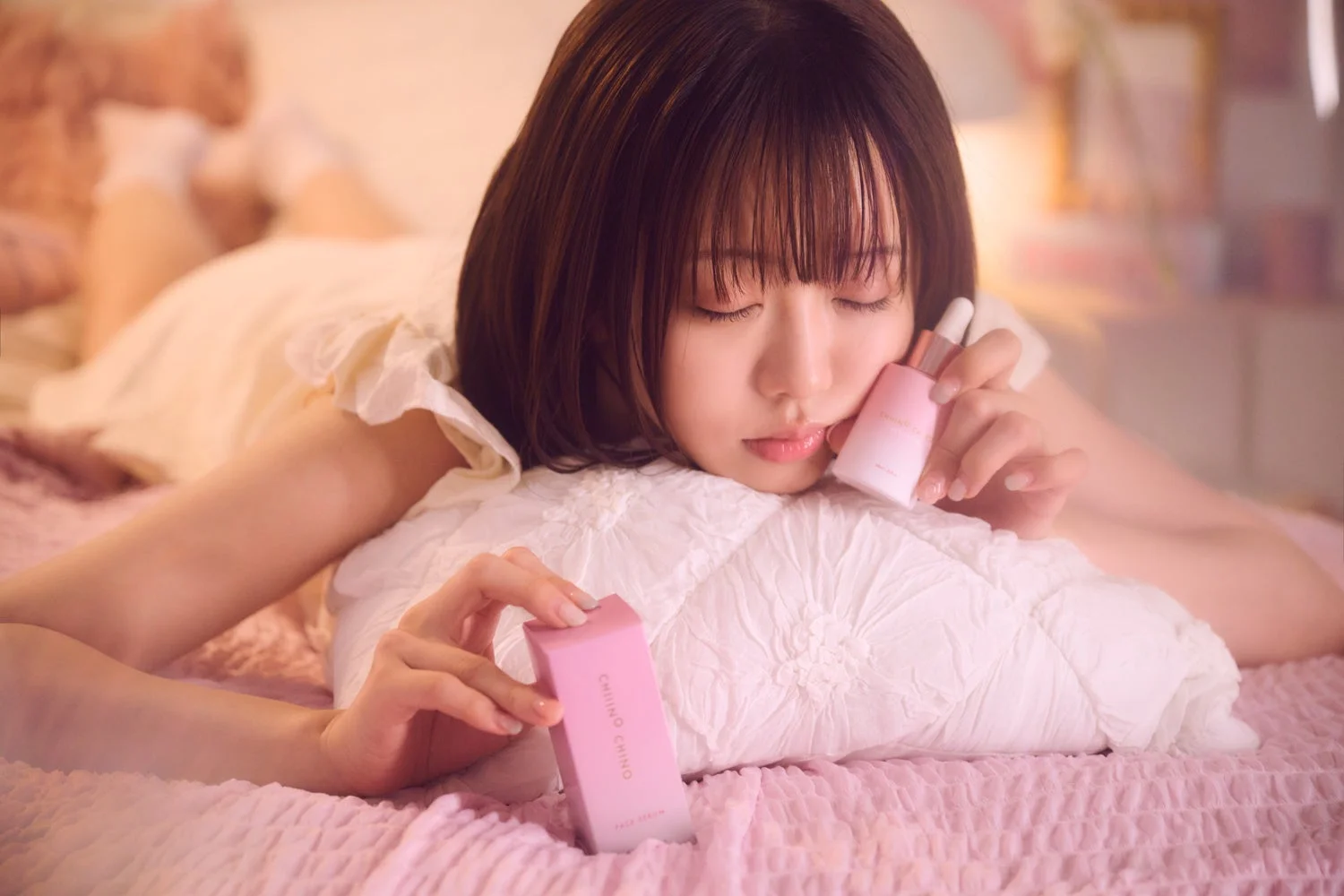 若い女性がベッドに横たわり、CHIINO CHIINOのフェイスセラムのボトルと箱を手に持っています。