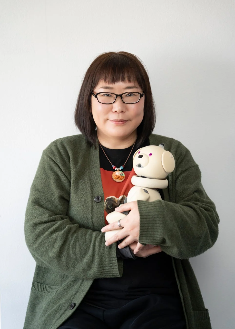 緑のカーディガンを着て眼鏡をかけた女性が、白い背景の前でクリーム色の犬型ロボット（AIBO）を抱きかかえているポートレート