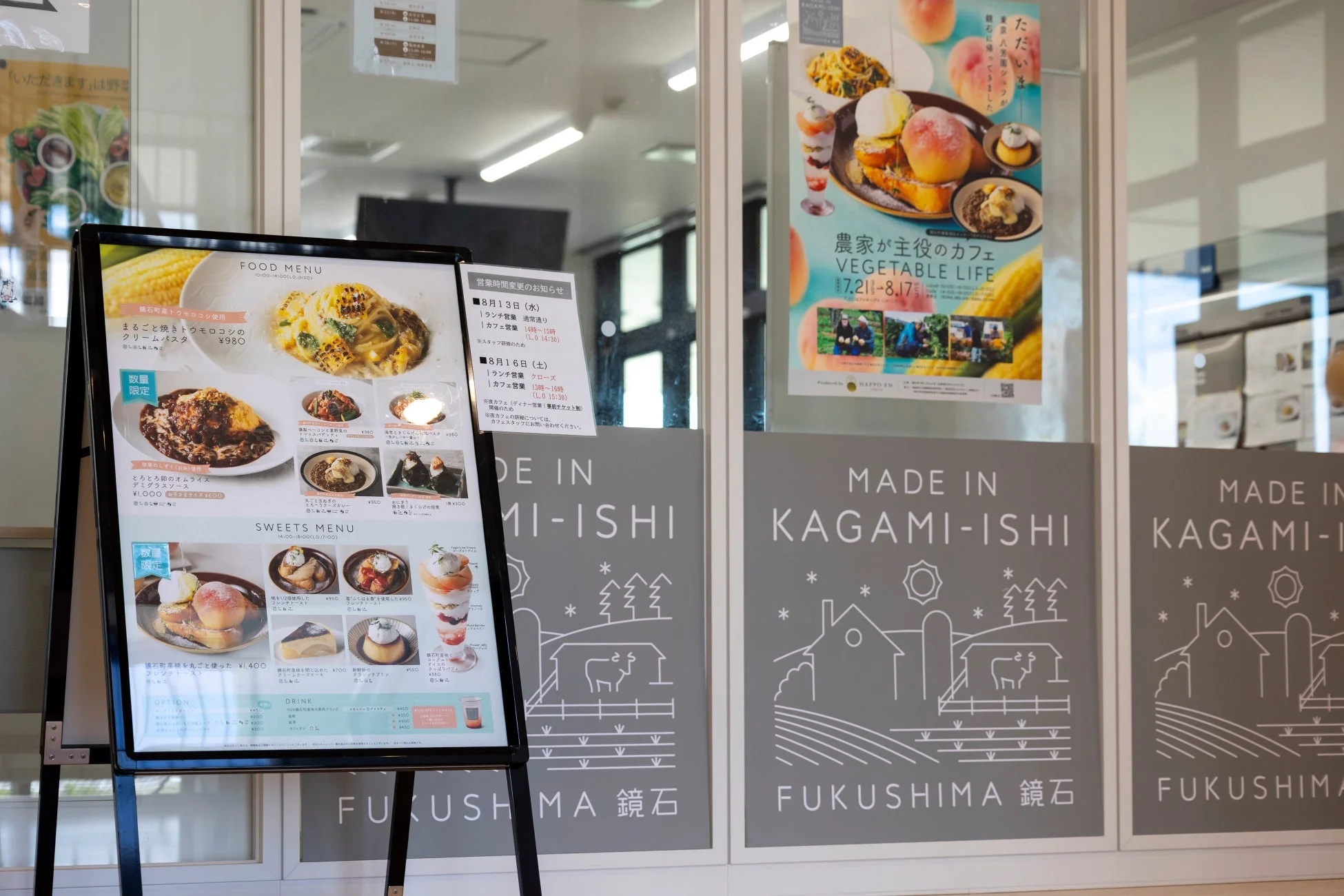 福島県鏡石町のカフェの店頭風景。フードメニューやスイーツ、営業時間変更の案内が掲示されており、地元産のトウモロコシや桃を使った料理やデザートが提供されていることがわかる