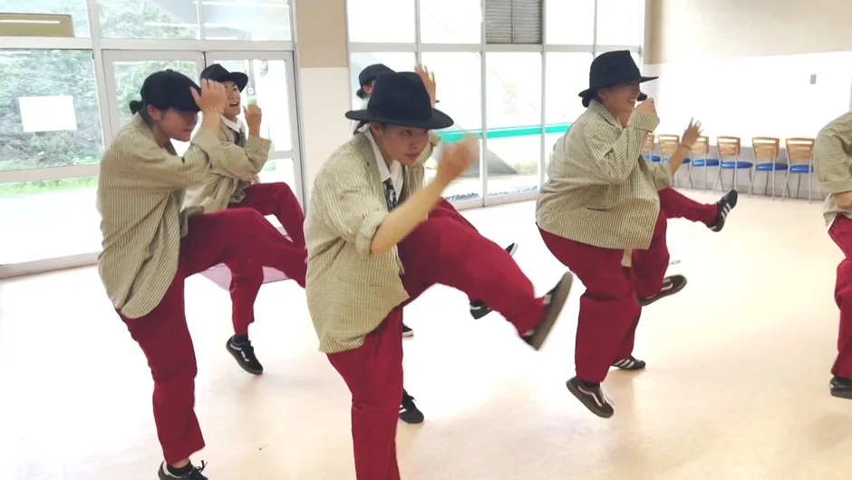 数人の若者が揃いのストライプシャツと赤いパンツ、帽子を身につけ、室内でダンスのパフォーマンスや練習をしている様子。