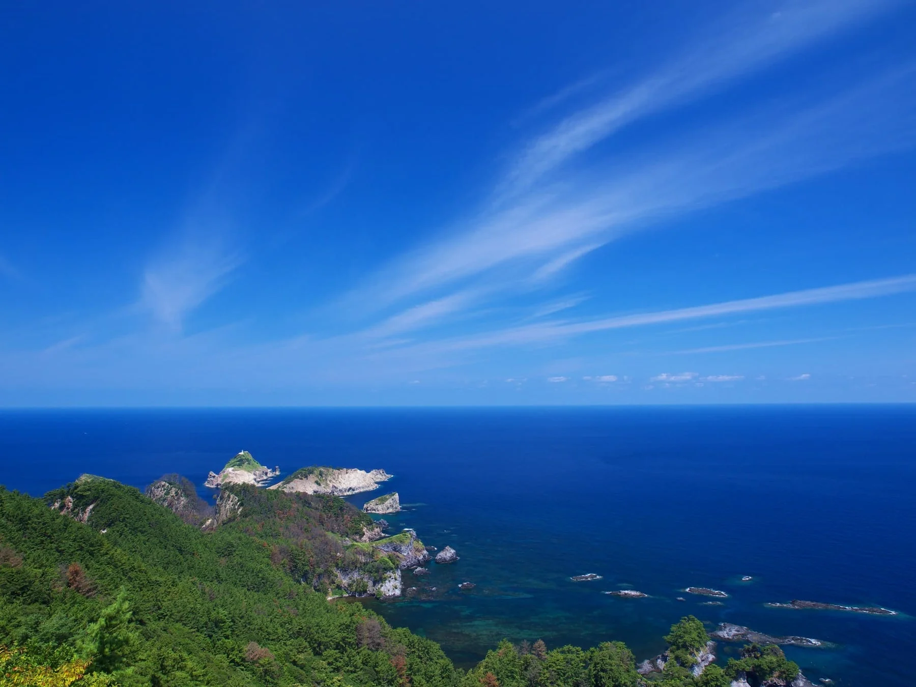 広大な青い海と青空が広がり、緑豊かな山々が連なる海岸線の絶景です。遠くには白い灯台が見える島々が点在し、爽やかな夏の自然風景が広がっています。